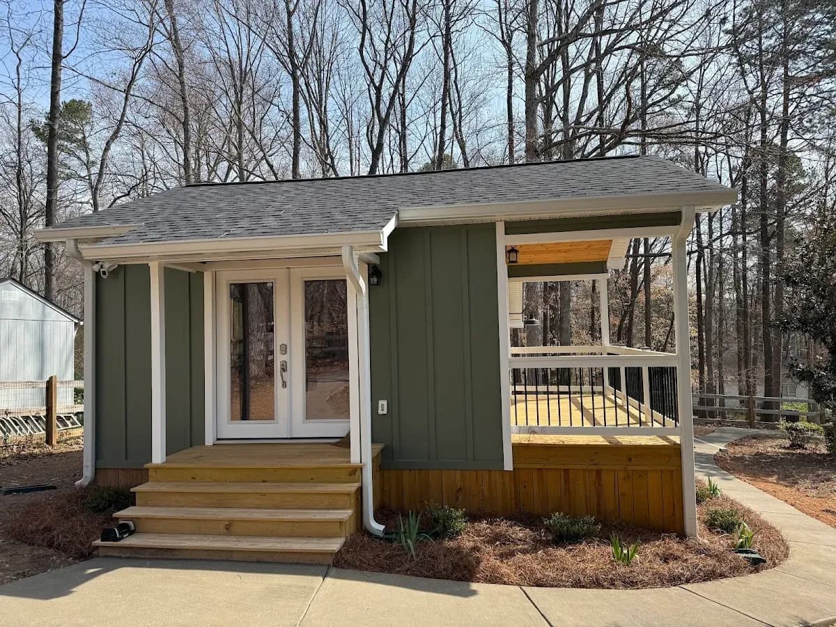 Custom She-Shed with Covered Porch by Alex's Lawnscape in Charlotte NC — Green board-and-batten outbuilding with covered side porch, French doors, wooden steps, and pine straw landscaping in a wooded setting