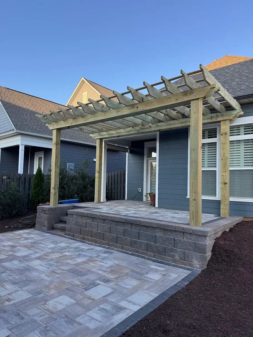 Pergola on Elevated Paver Patio by Alex's Lawnscape in Charlotte NC — Timber pergola over an elevated paver patio with block retaining wall steps and fresh mulch beds at a blue-sided home