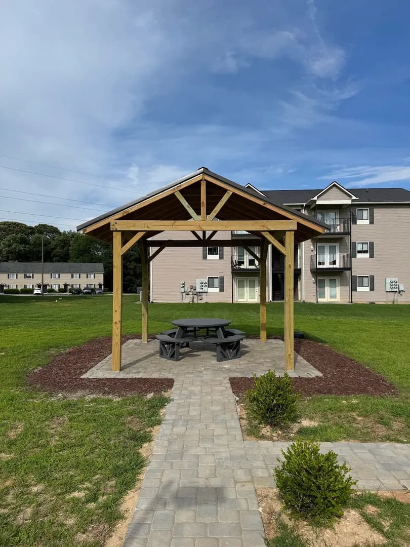 Pavilion & Picnic Area by Alex's Lawnscape in Charlotte NC — Open-gable timber pavilion with metal roof over a paver pad and picnic table, connected by paver walkway with boxwood plantings