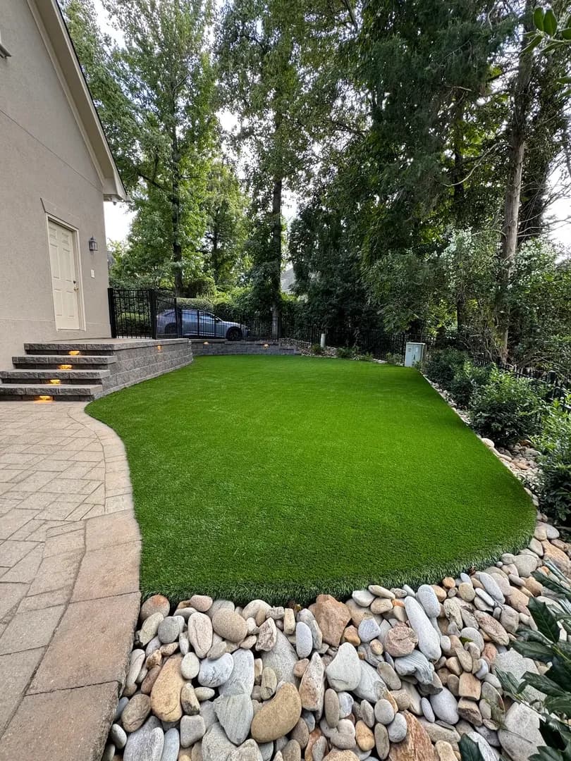 Artificial Turf with Paver Patio by Alex's Lawnscape in Charlotte NC — Lush synthetic turf lawn edged with river rock and pavers, retaining wall with step lights, in a shaded backyard