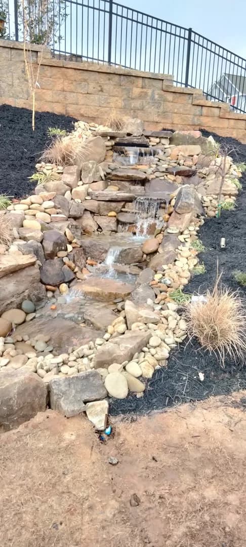 Cascading Rock Water Feature by Alex's Lawnscape in Charlotte NC — Multi-tier cascading water feature built with stacked natural stone and river rock below a retaining wall with fresh mulch beds
