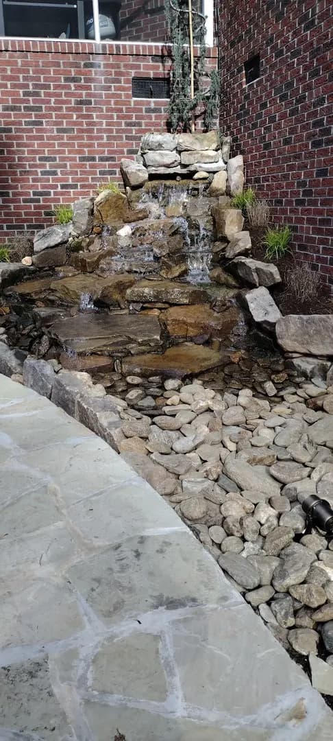 Natural Stone Water Feature by Alex's Lawnscape in Charlotte NC — Pondless waterfall built with stacked natural stone and river rock, with accent plantings and a flagstone border